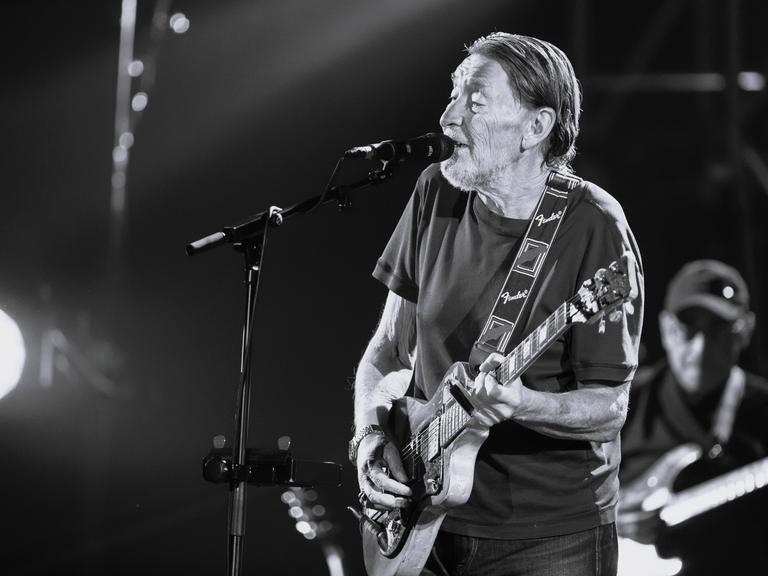 Chris Rea steht auf der Bühne. Er spielt Gitarre und singt in ein Mikrofon. Das Bild ist schwarz-weiß. (Quelle: picture alliance)