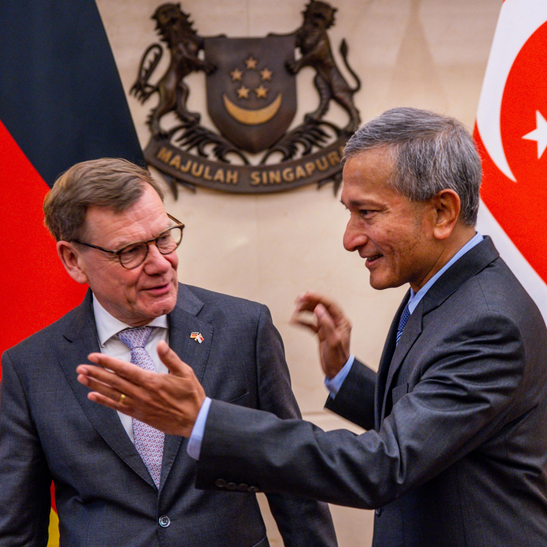 Die beiden stehen lächelnn in einem Raum nebeneinander. Balakrishnan weist Wadephul den Weg nach rechts. Dahinter die Flaggen beider Länder vor einer Wand. 