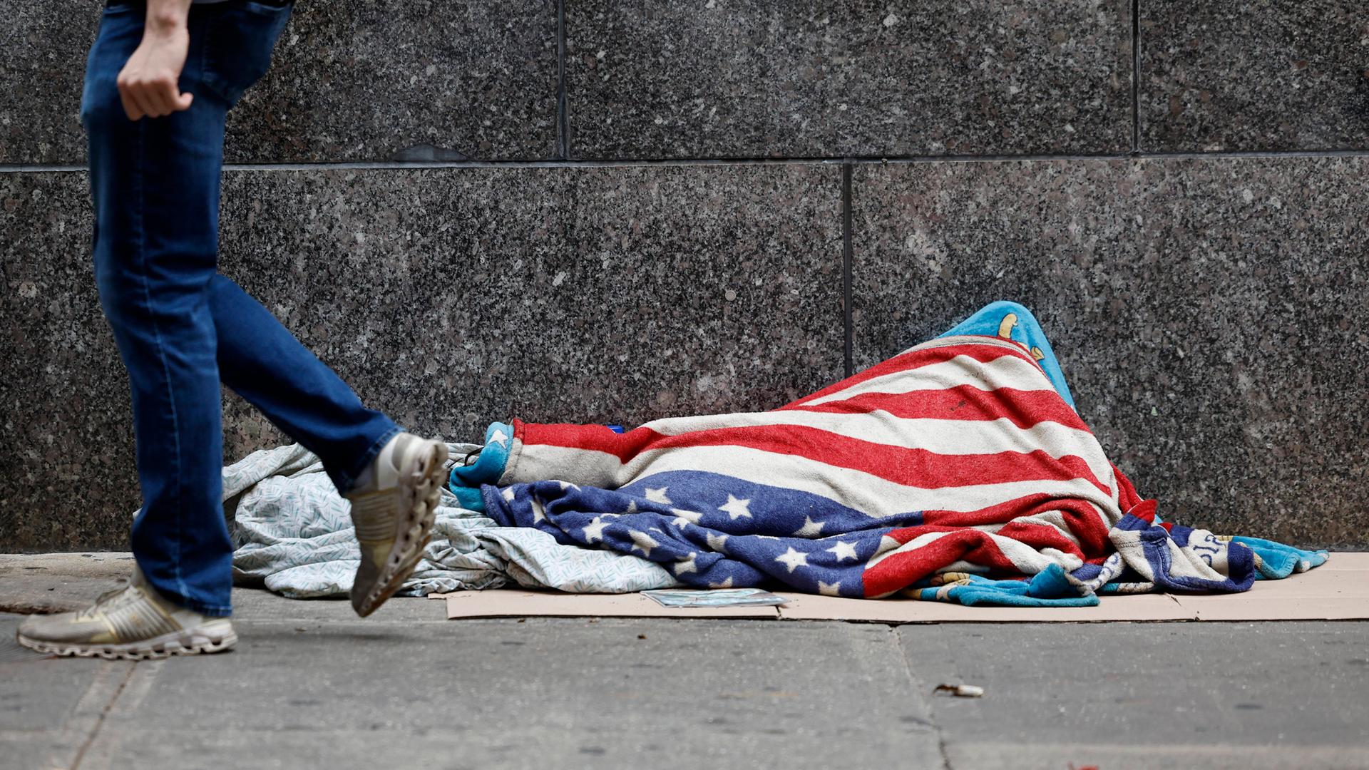 Vermutlich eine obachlose Person mit einer US-Flagge bedeckt, liegt auf einer Straße nahe der Wall Street. Aufgenommen am 18.6.2025 in New York City. Vermutlich eine obachlose Person mit einer US-Flagge bedeckt, liegt auf einer Straße nahe der Wall Street. Aufgenommen am 18.6.2025 in New York City.