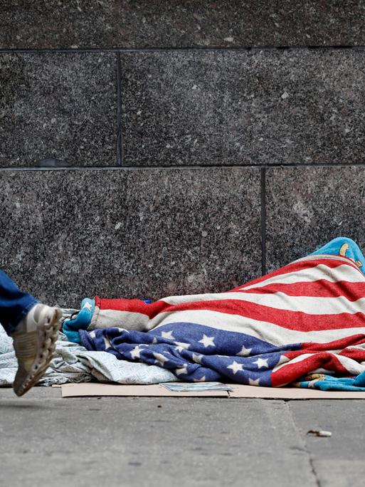 Vermutlich eine obachlose Person mit einer US-Flagge bedeckt, liegt auf einer Straße nahe der Wall Street. Aufgenommen am 18.6.2025 in New York City. 