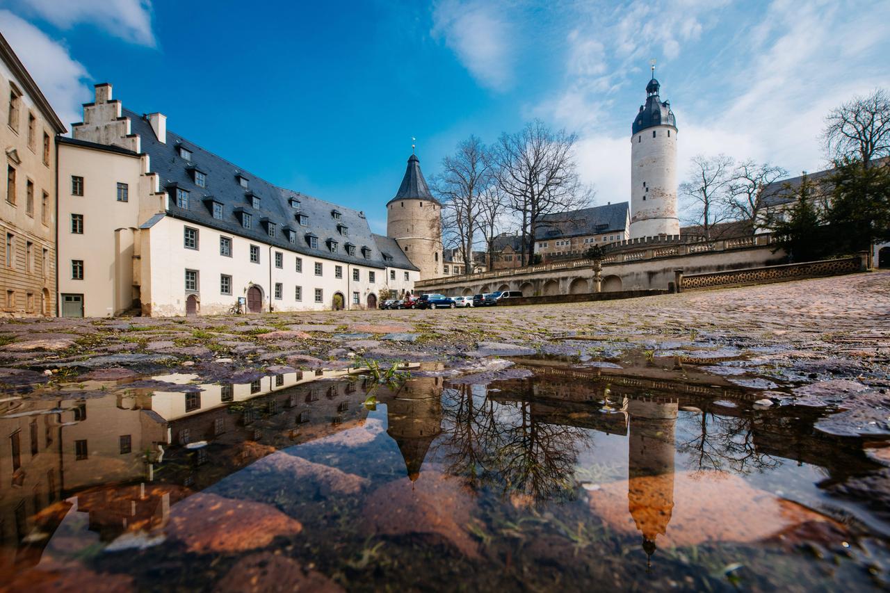 Das Bild zeigt eine Außenansicht des alten Residenzschlosses Altenburg. Im Hintergrund ist das helle Gebäude zu sehen, im Vordergrund, aus einer Weitwinkelperspektive, eine spiegelnde Wasserpfütze. Der Platz ist menschenleer.