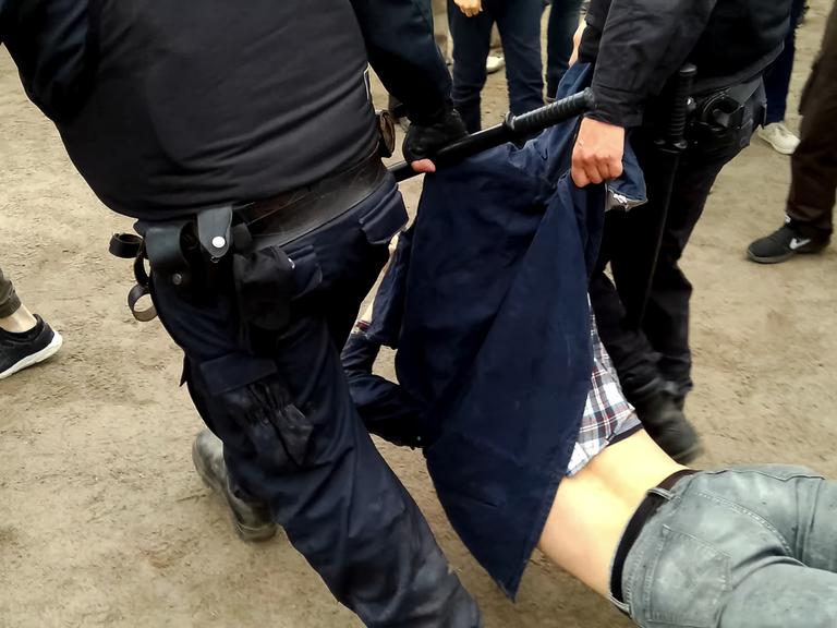 Zwei Sicherheitsbeamte mit Schlagstöcken und in schwarzen Uniformen tragen brutal eine Person weg.