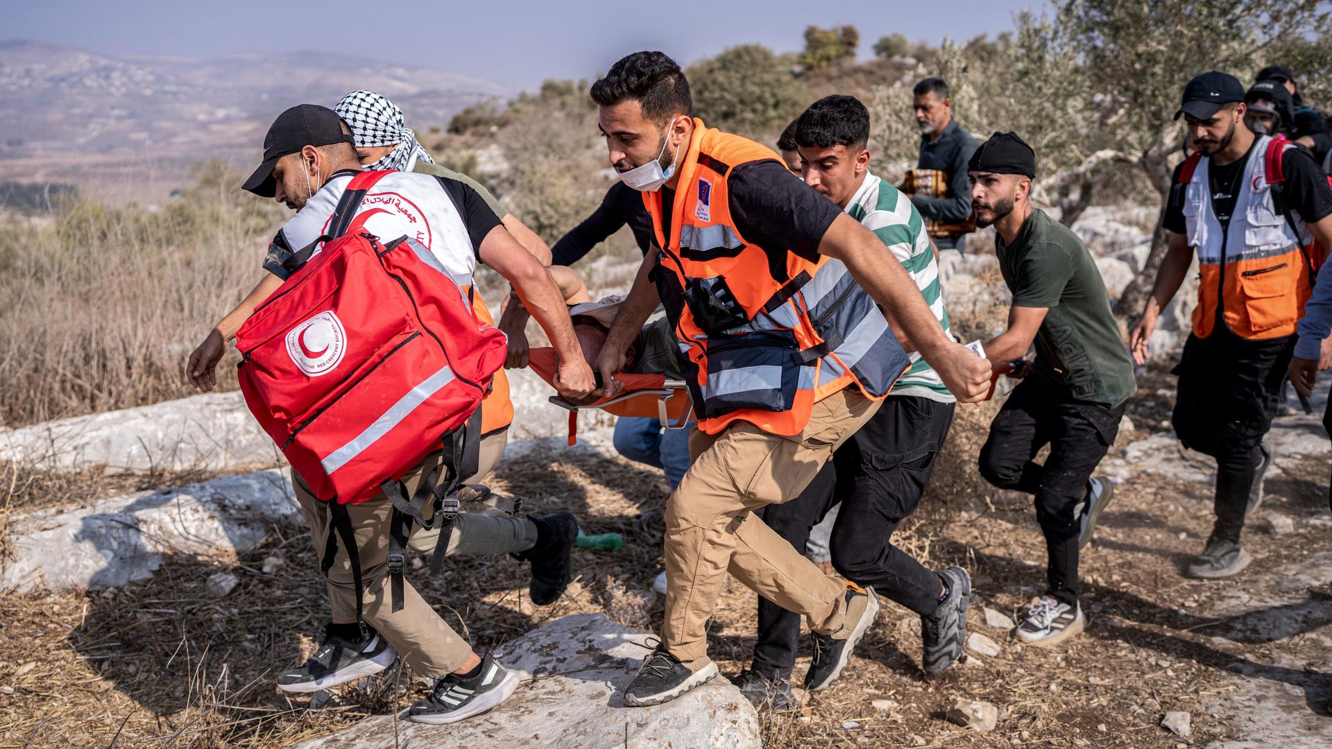 Mehrere Männer tragen den Verletzte im Laufschritt in einer Bahren an Olivenbäumen vorbei. Einige von ihnen tragen leuchtorangene Warnwesten. Mehrere Männer tragen den Verletzte im Laufschritt in einer Bahren an Olivenbäumen vorbei. Einige von ihnen tragen leuchtorangene Warnwesten.