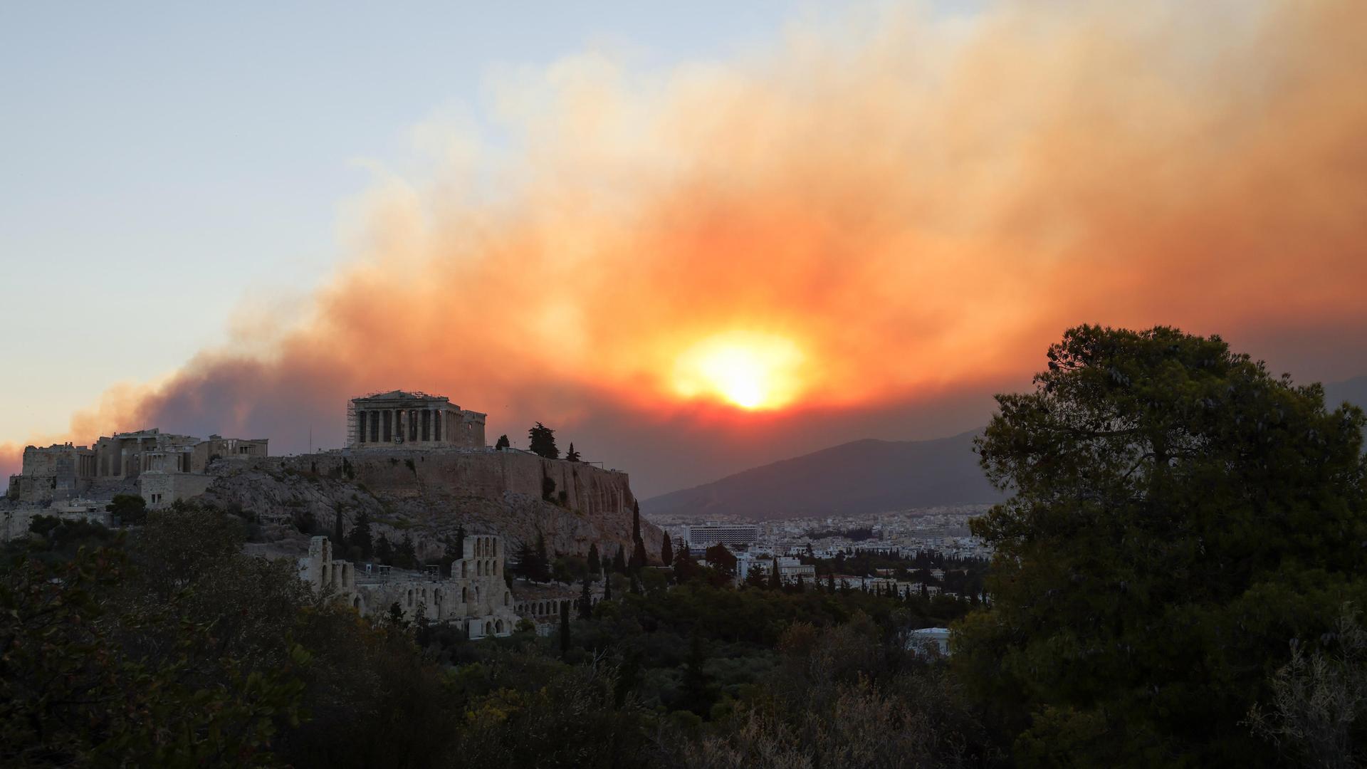 Hinter der Akropolis von Athen steigt Rauch und oranges Licht von den nahegelegenen Waldbränden auf.