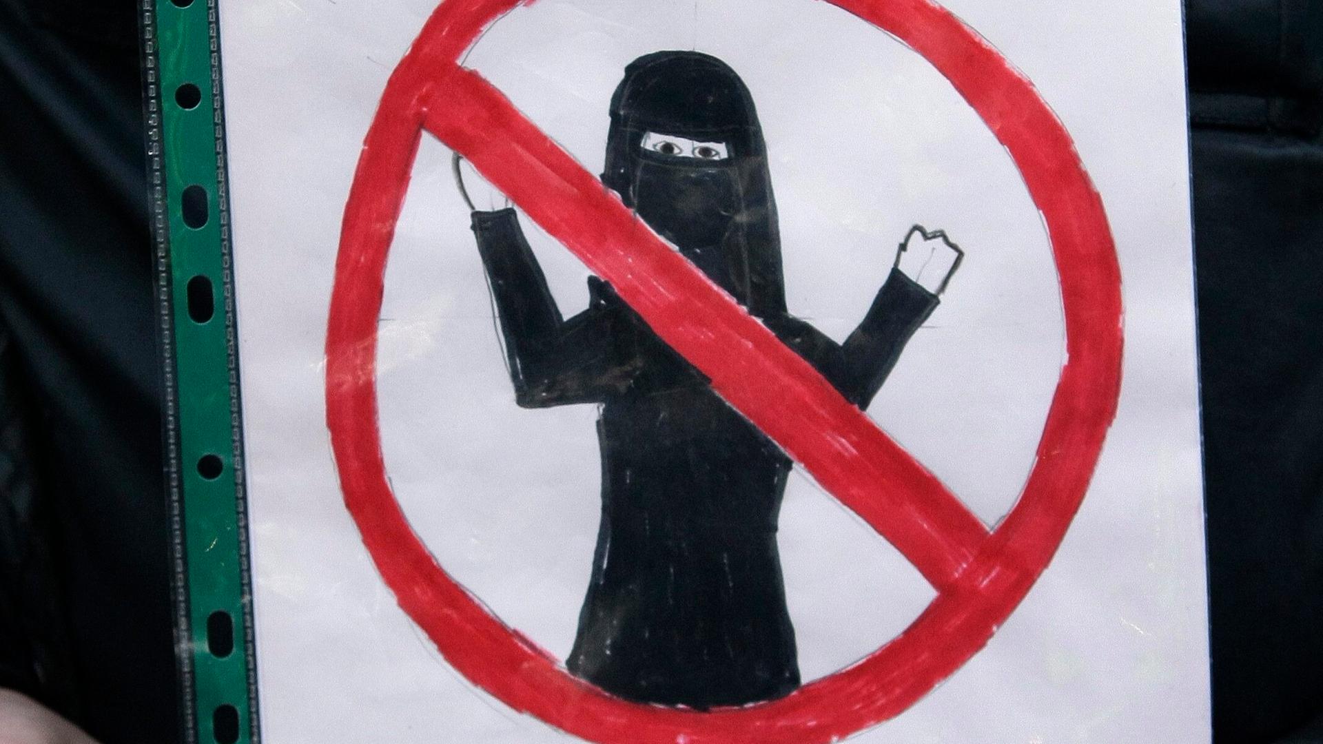 Jemand hält ein DIN A4-Blatt in einer Klarsichthülle. Auf das Papier ist mit Buntstiften ein rotes Verbotszeichen gemalt mit einer verschleierten Frau in der Mitte. 