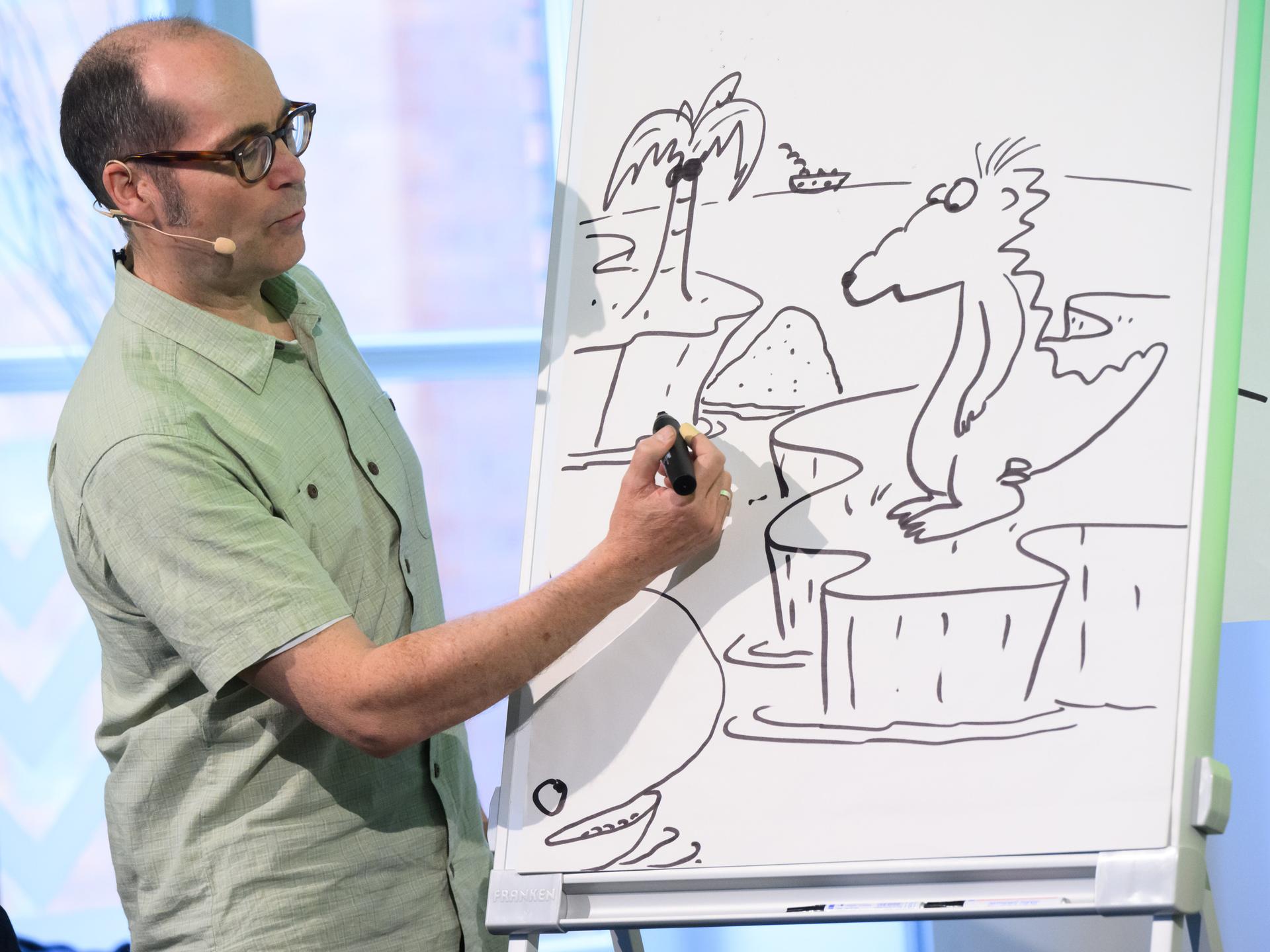 Kinderbuchautor Ingo Siegner zeigt, wie er seine Buchfigur Drache Kokosnuss zeichnet. Er steht vor einem Zeichenblock und zeichnet. Kinderbuchautor Ingo Siegner zeigt, wie er seine Buchfigur Drache Kokosnuss zeichnet. Er steht vor einem Zeichenblock und zeichnet.