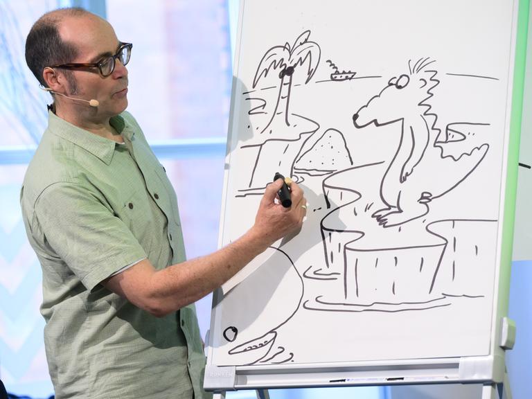 Kinderbuchautor Ingo Siegner zeigt, wie er seine Buchfigur Drache Kokosnuss zeichnet. Er steht vor einem Zeichenblock und zeichnet. Kinderbuchautor Ingo Siegner zeigt, wie er seine Buchfigur Drache Kokosnuss zeichnet. Er steht vor einem Zeichenblock und zeichnet.