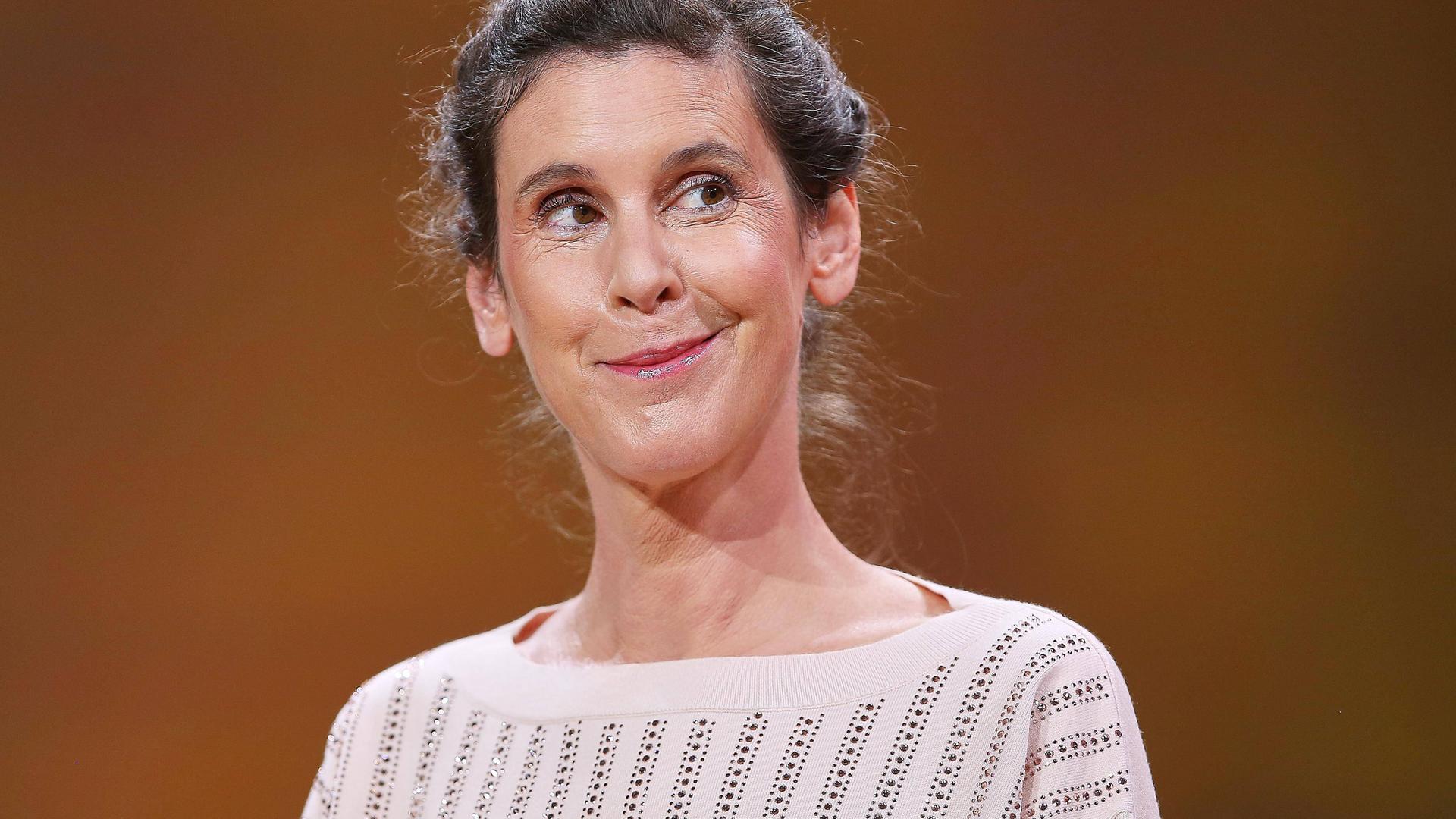 Daniela Dröscher steht vor einem neutralen Hintergrund, lächelt verschmitzt und blickt dabei ins Off