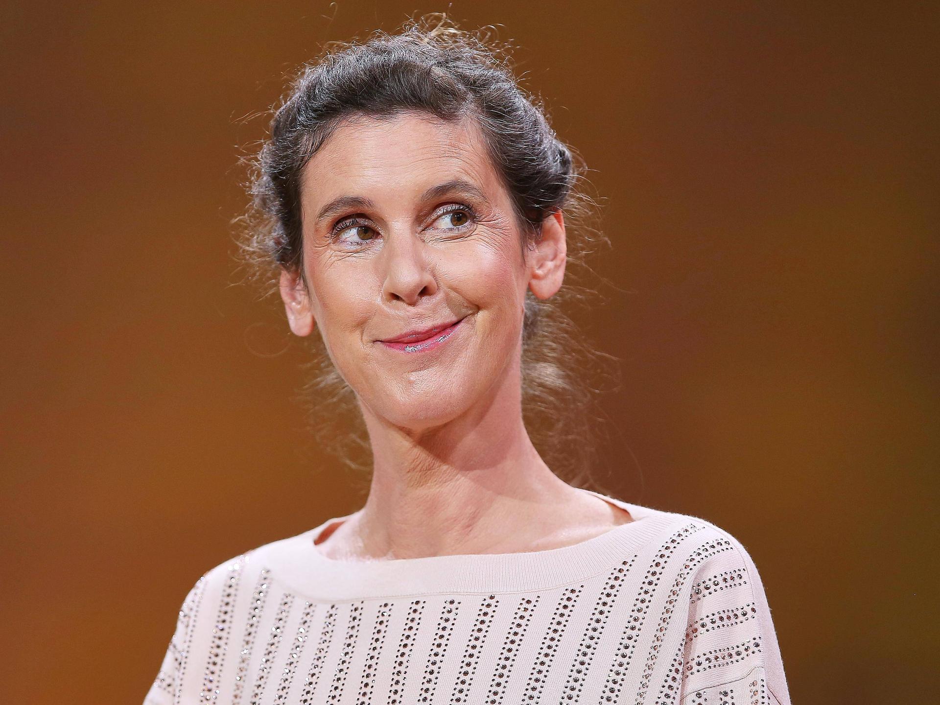 Daniela Dröscher steht vor einem neutralen Hintergrund, lächelt verschmitzt und blickt dabei ins Off