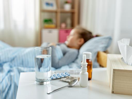 Ein krankes Kind liegt im Bett, im Vordergrund stehen Hustensaft, Nasenspray, Tabletten und ein Glas Wasser.