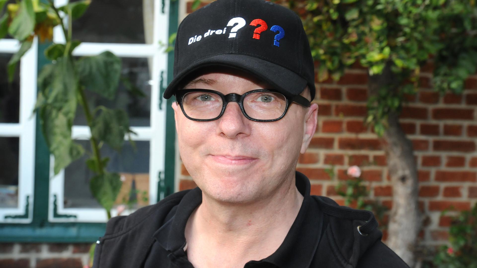 Schauspieler Oliver Rohrbeck mit einer Mütze mit dem Logo der Hörspielserie drei Fragezeichen.
