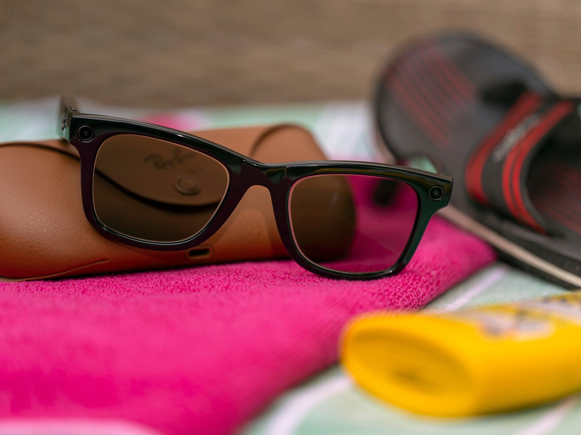 Eine Smart Glasses liegt auf einem Strandhandtuch zwischen Sonnencreme und Badelatschen.