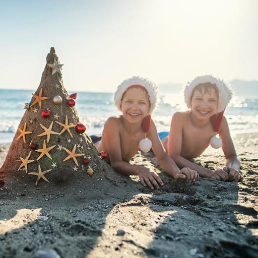 Drei Kinder liegen am Strand mit Weihnachtsmannmützen und einer als Weihnachtsbaum dekorierten Sandburg