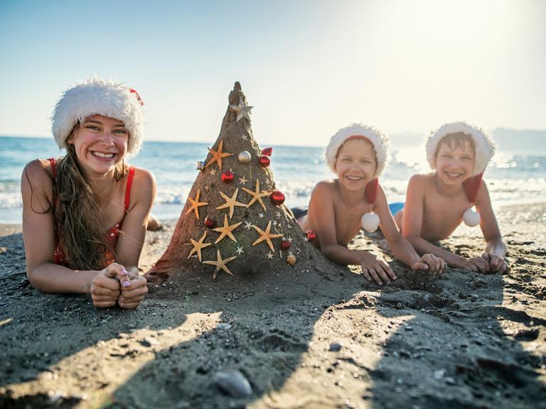 Drei Kinder liegen am Strand mit Weihnachtsmannmützen und einer als Weihnachtsbaum dekorierten Sandburg