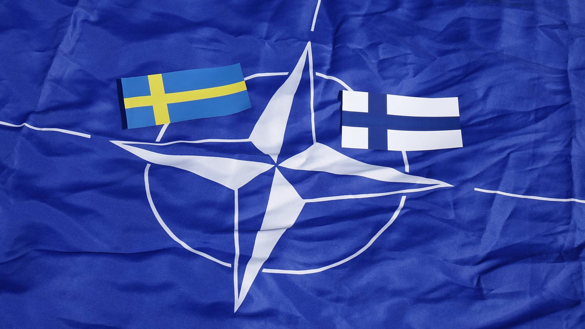 Kleine Flaggen von Schweden nund Finnland laigen auf einer großen Flagge der mit dem NATO-Logo. 