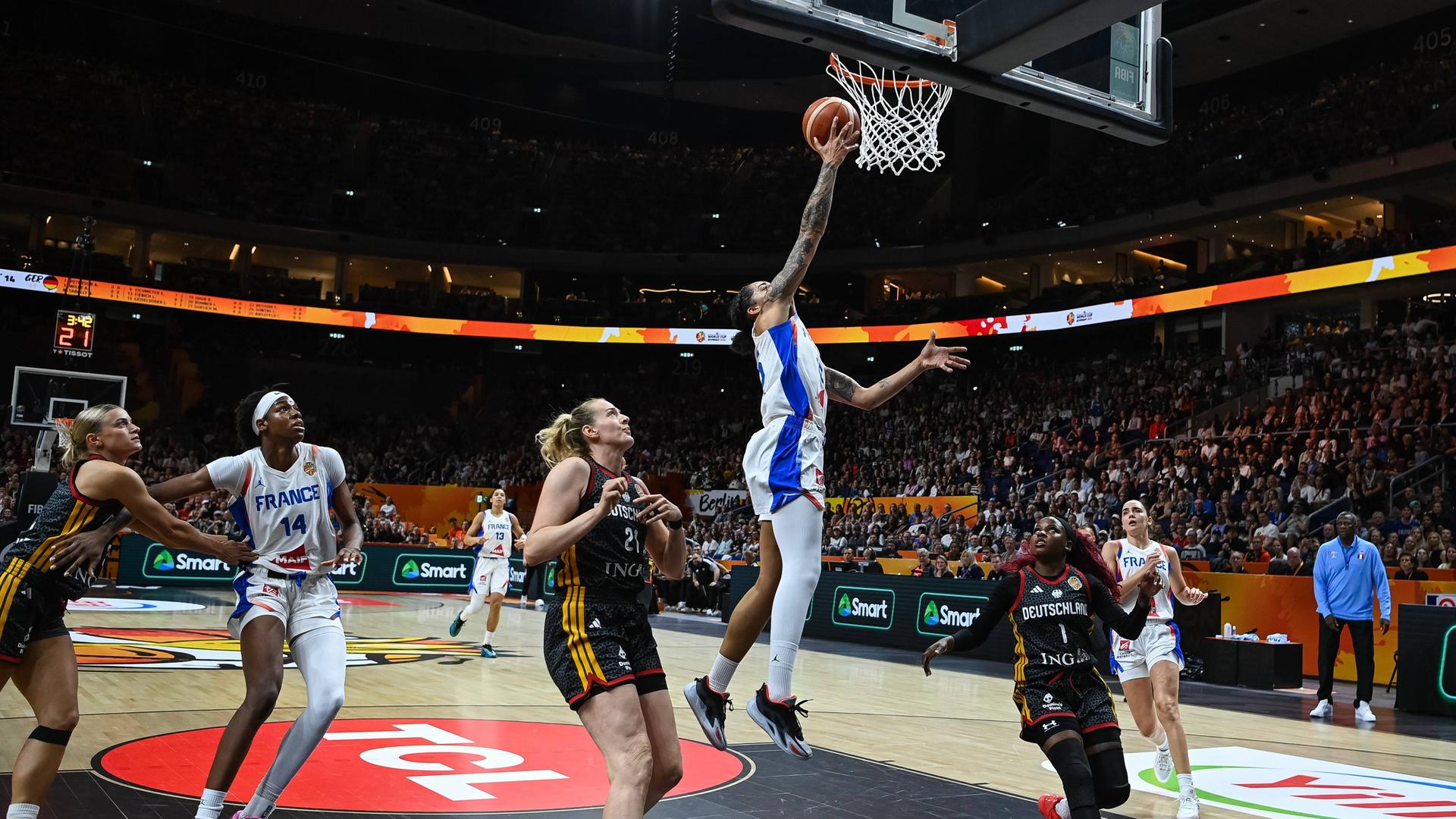 Eine französische Basketballerin springt mit dem Ball in der Hand zum Korb. Andere französische und deutsche Spielerinnen schauen ihr zu, ohne eingreifen zu können. 