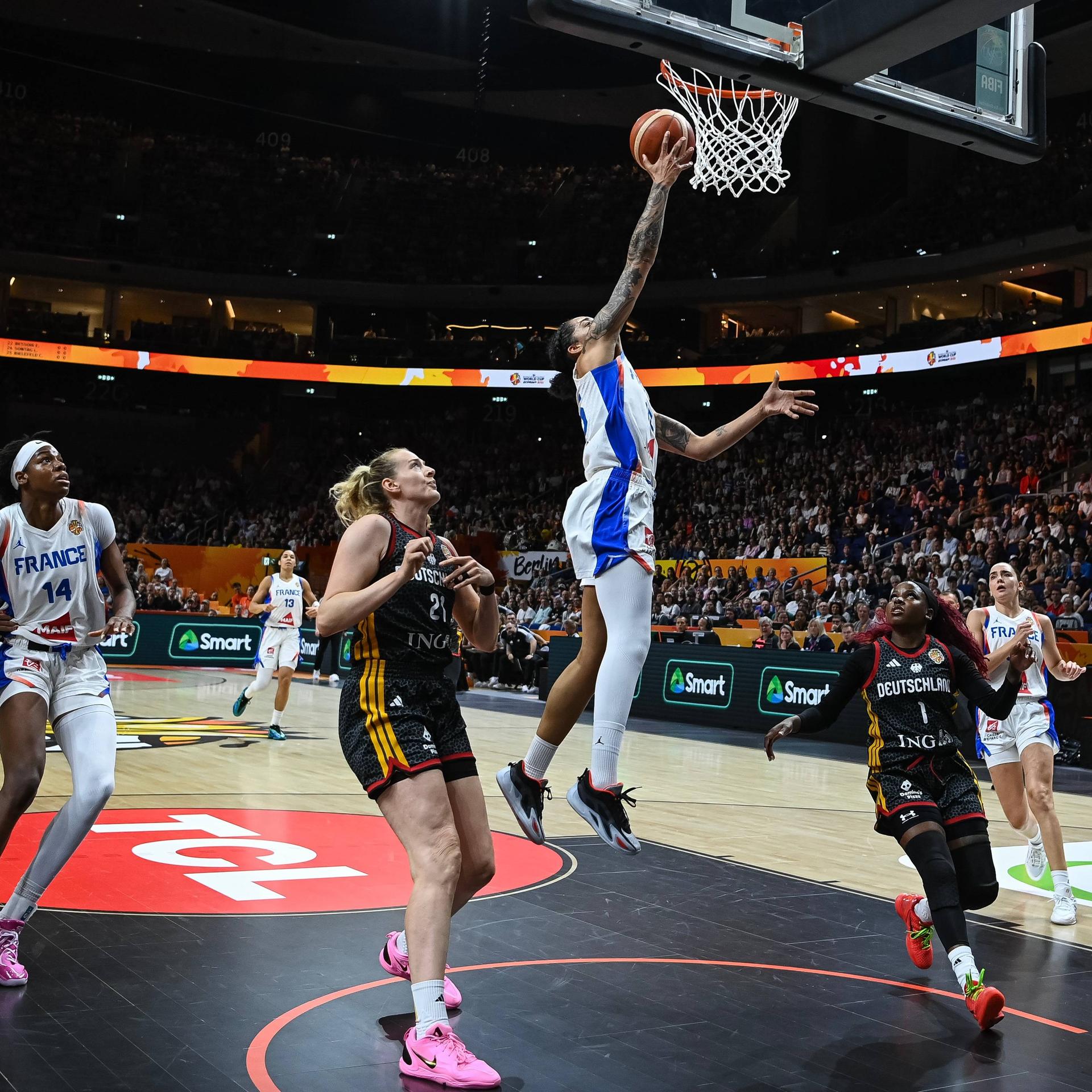 Eine französische Basketballerin springt mit dem Ball in der Hand zum Korb. Andere französische und deutsche Spielerinnen schauen ihr zu, ohne eingreifen zu können. 