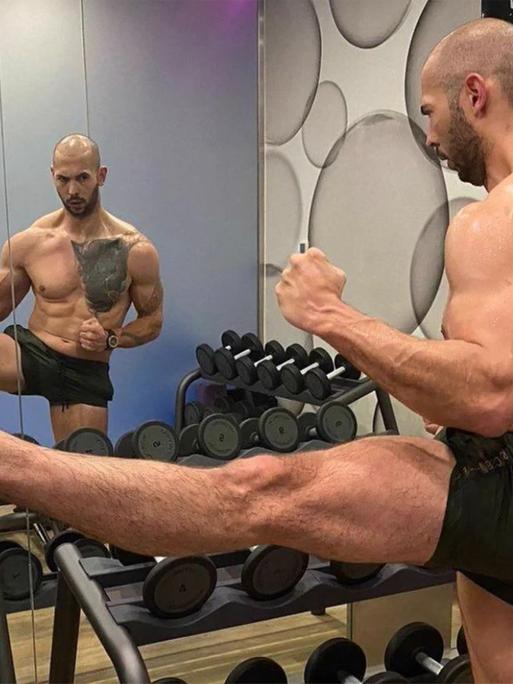 Andrew Tate in knapper kurzer Hose im Gym, er flext seine Muskeln und betrachtet sich im Spiegel.