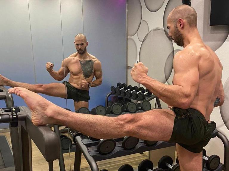 Andrew Tate in knapper kurzer Hose im Gym, er flext seine Muskeln und betrachtet sich im Spiegel.
