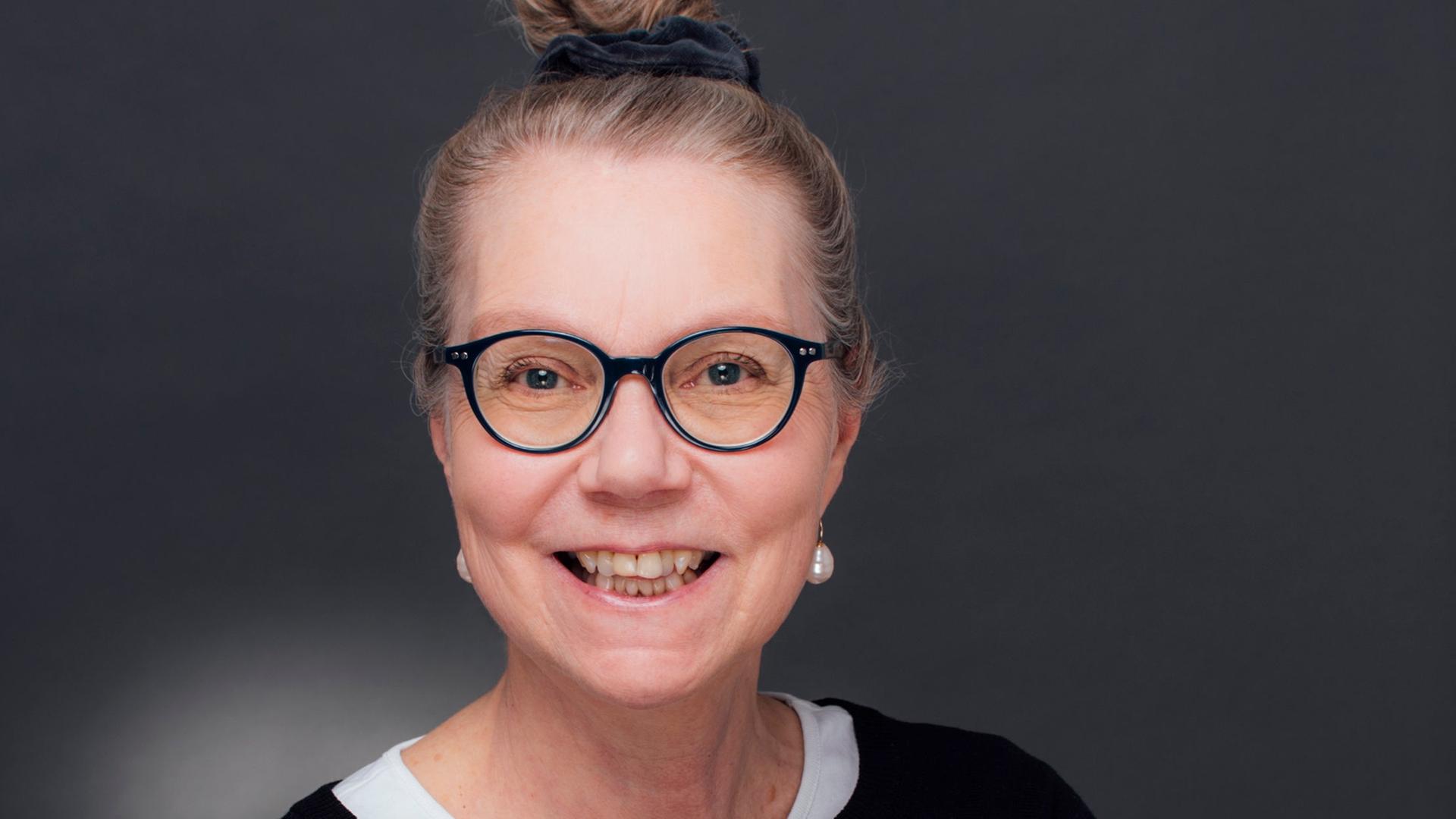 Ursula Gräfe steht mit verschränkten Armen in einem Fotostudio vor grauem Hintergrund. Sie trägt eine schwarz umrandete Brille und lacht in die Kamera. Ursula Gräfe steht mit verschränkten Armen in einem Fotostudio vor grauem Hintergrund. Sie trägt eine schwarz umrandete Brille und lacht in die Kamera.