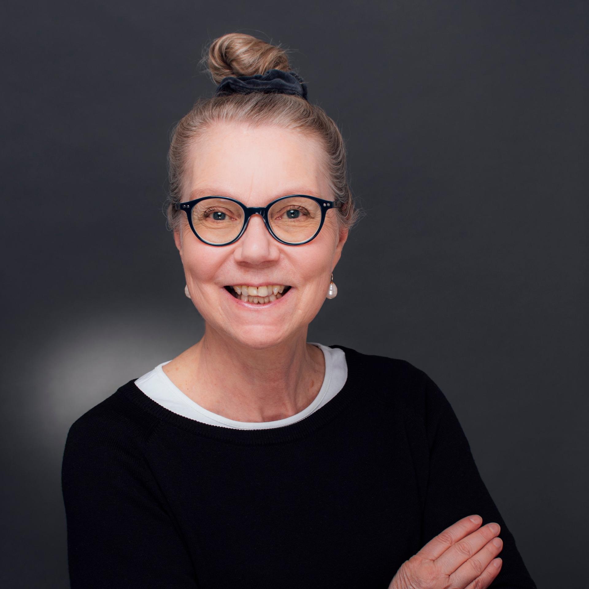 Ursula Gräfe steht mit verschränkten Armen in einem Fotostudio vor grauem Hintergrund. Sie trägt eine schwarz umrandete Brille und lacht in die Kamera.
