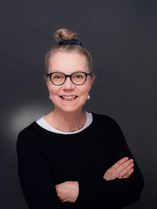 Ursula Gräfe steht mit verschränkten Armen in einem Fotostudio vor grauem Hintergrund. Sie trägt eine schwarz umrandete Brille und lacht in die Kamera.