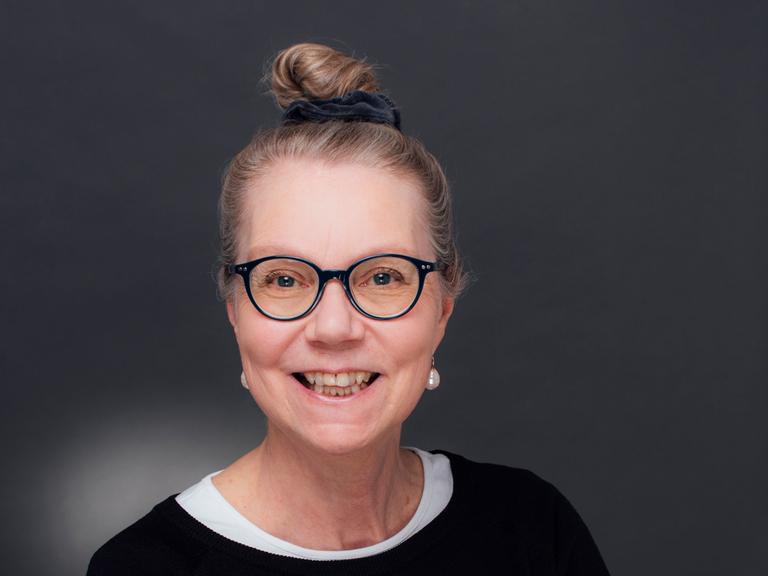 Ursula Gräfe steht mit verschränkten Armen in einem Fotostudio vor grauem Hintergrund. Sie trägt eine schwarz umrandete Brille und lacht in die Kamera.