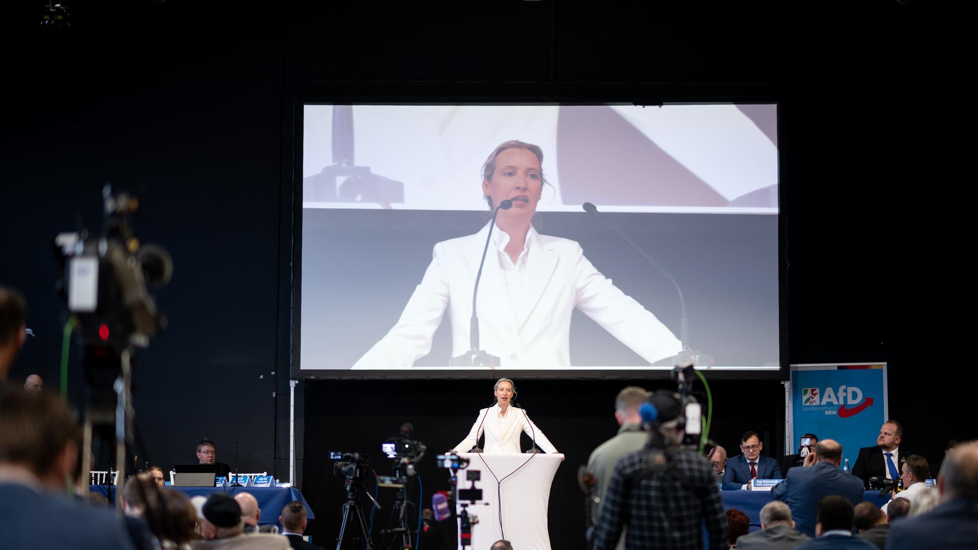Alice Weidel, Vorsitzende der AfD-Bundestagsfraktion, spricht beim Landesparteitag der AfD in NRW. Sie steht im weißen Anzug auf einer Bühne. Auf einem großen Bildschirm hinter ihr wird ihr Auftritt übertragen.