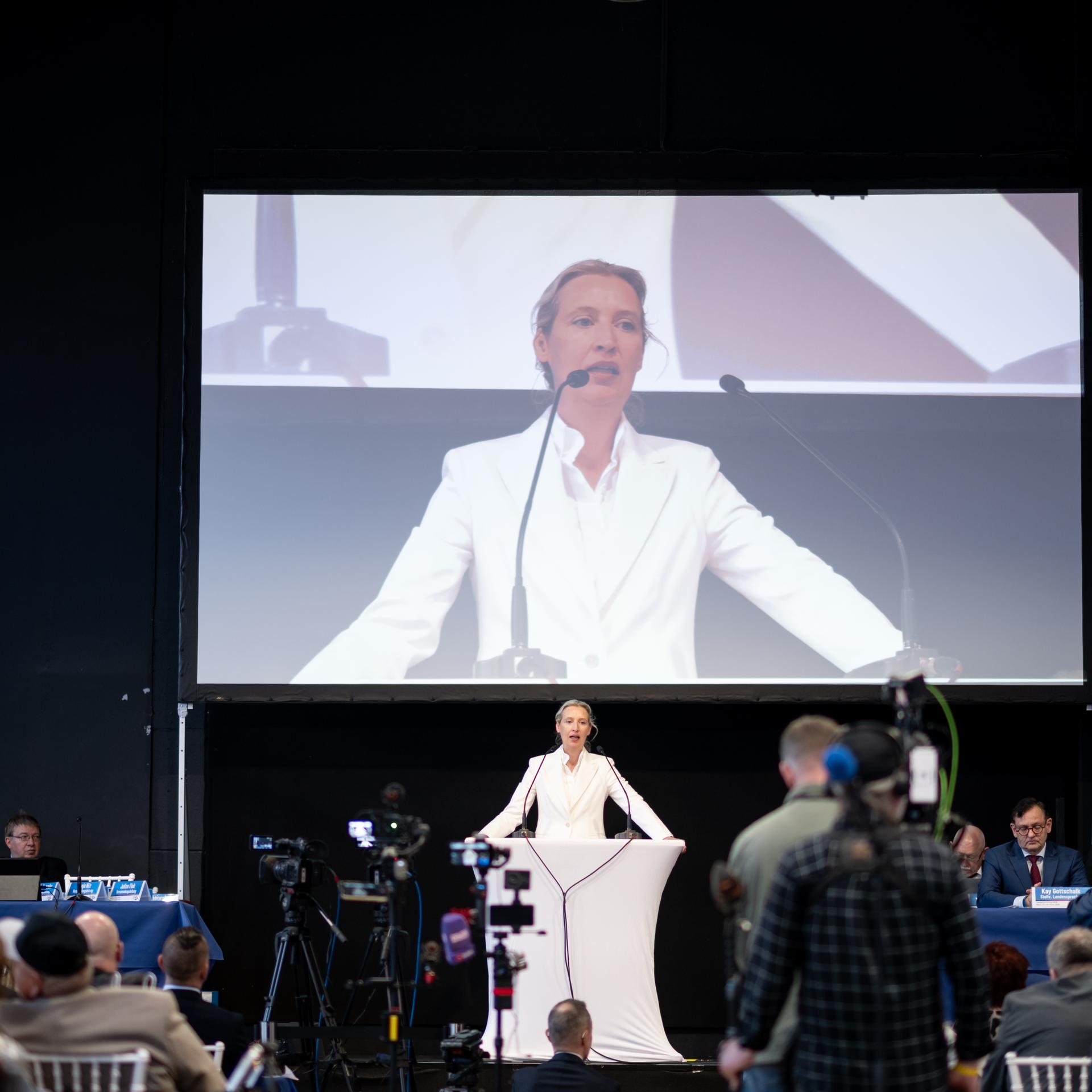 Alice Weidel, Vorsitzende der AfD-Bundestagsfraktion, spricht beim Landesparteitag der AfD in NRW. Sie steht im weißen Anzug auf einer Bühne. Auf einm großen Bildschirm hinter ihr, wird ihr Auftritt übertragen.