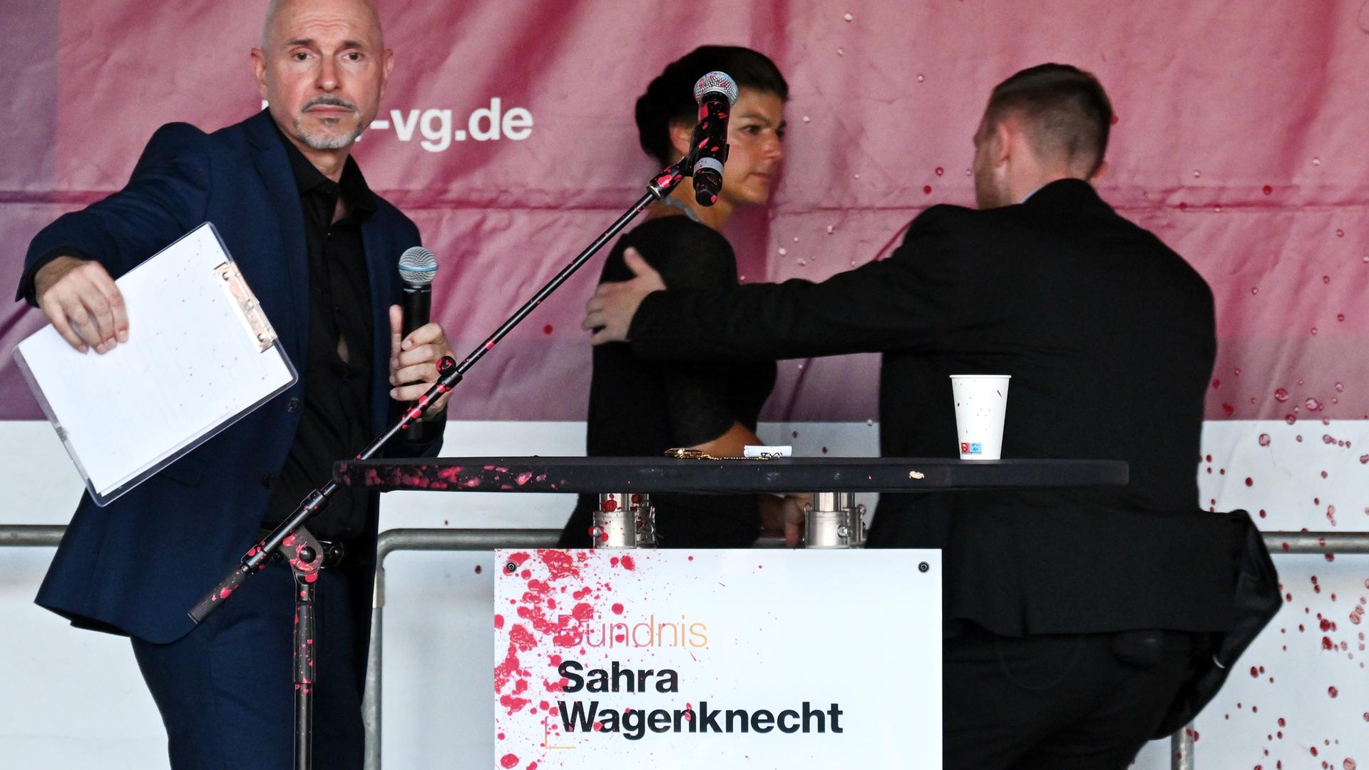 Hinter einem Rednerpult, das rote Farbspritzer hat, wird Wagenknecht von der Bühne begleitet