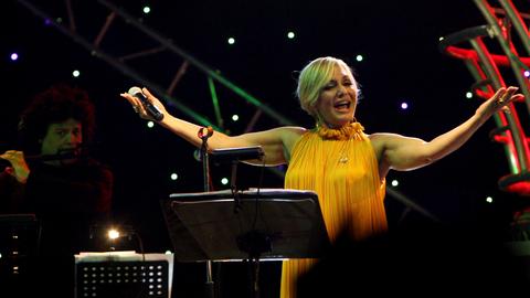 Die iranische Sängerin Googoosh steht singend auf einer Bühne und breitet die Arme aus.