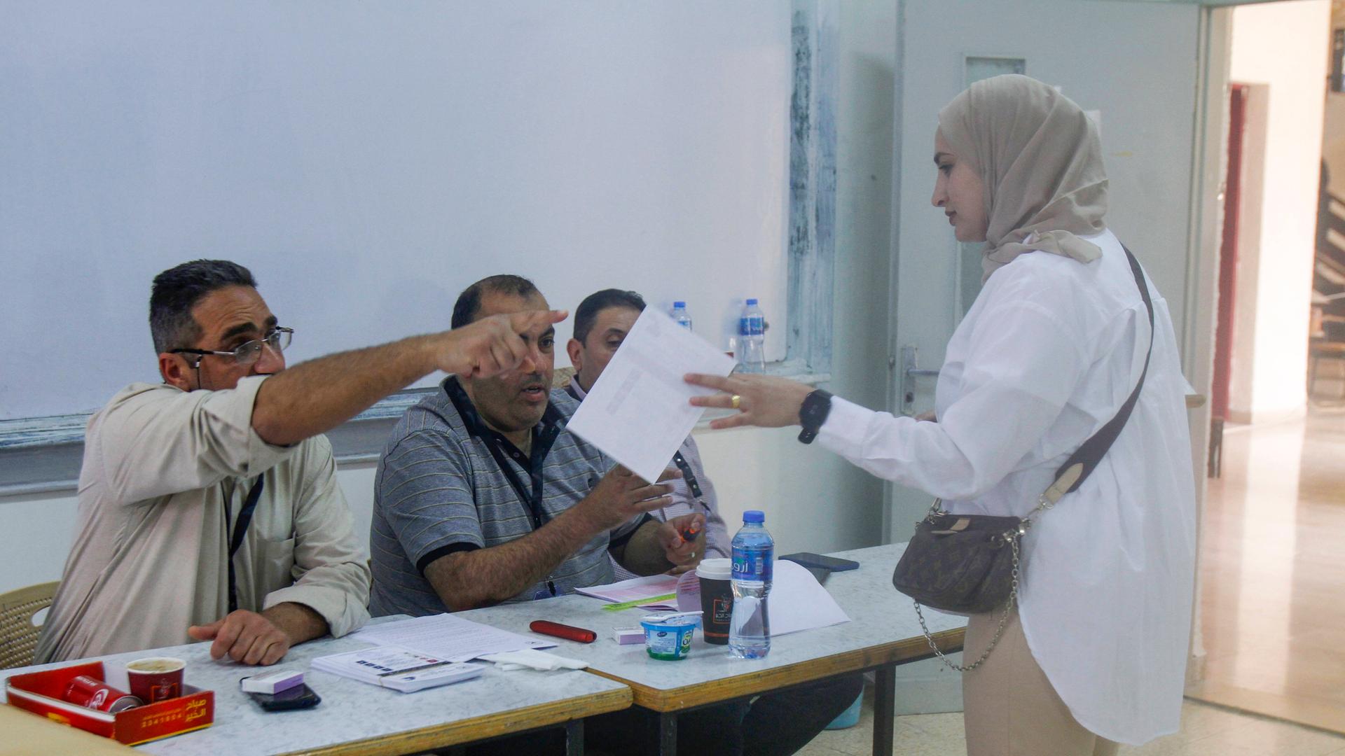 Eine junge Frau mit Kopftuch im Wahlbüro. Sie hat einen Stimmzettel in der Hand. Ein Mann weist ihr den Weg.