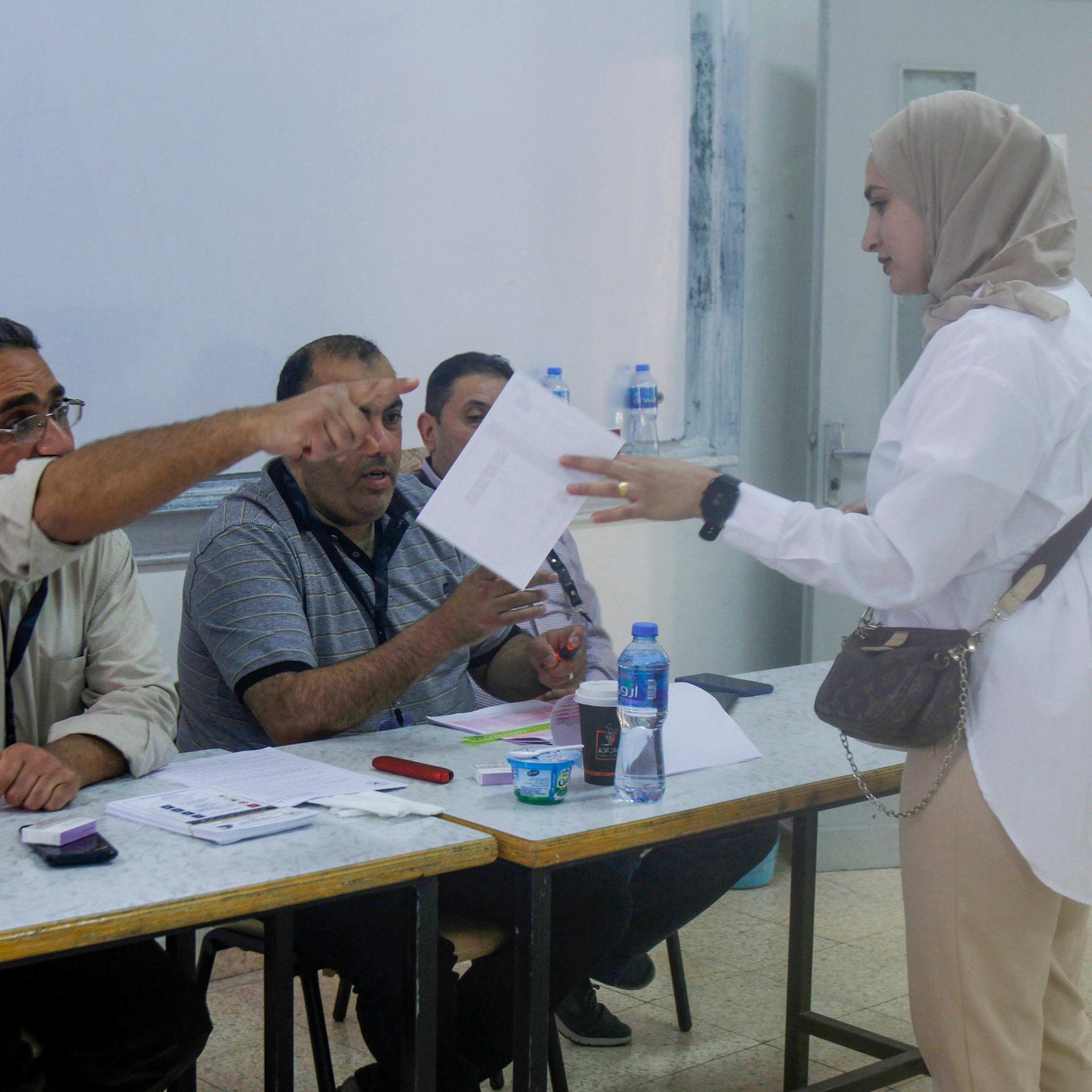Eine junge Frau mit Kopftuch im Wahlbüro. Sie hat einen Stimmzettel in der Hand. Ein Mann weist ihr den Weg.
