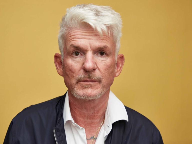 Der Schriftsteller Heinz Strunk blickt vor gelbem Hintergrund ernst in die Kamera