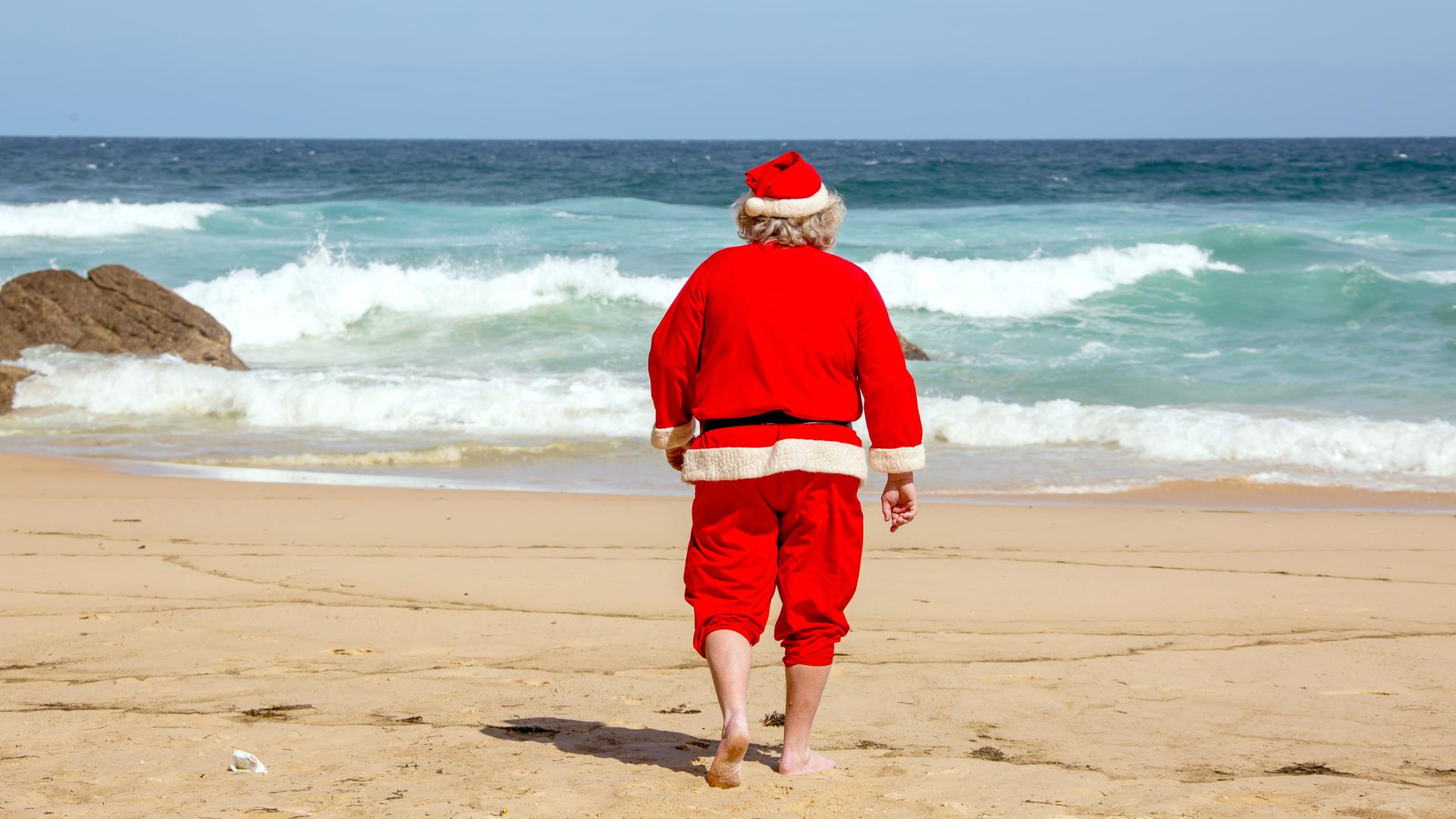 Ein Weihnachtsmann läuft barfuß am Strand direkt auf das blaue Meer zu.