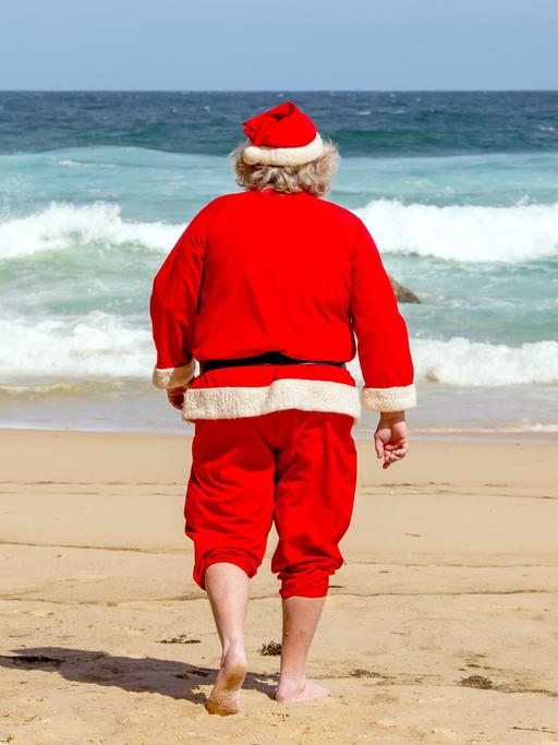 Ein Weihnachtsmann läuft barfuß am Strand direkt auf das blaue Meer zu.
