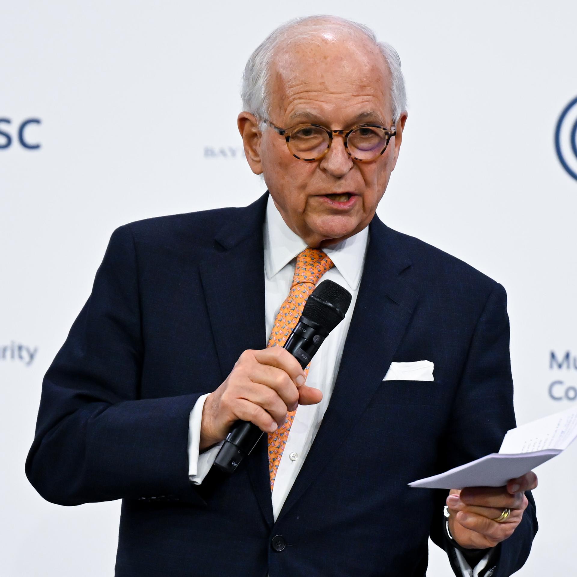 Ischinger steht auf der Bühne, spricht ins Mikrofon und hält mit der anderen Hand einen Zettel. Im Hintergrund eine weiße Wand mit den Schriftzügen und Logos der Konferenz.