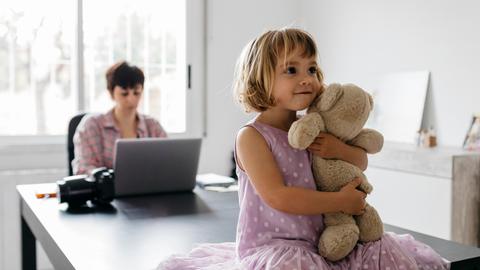Eine Frau arbeitet an einem Tisch mit dem Laptop. Auf dem Tisch sitzt ein Mädchen, das einen Teddybären im Arm hält.