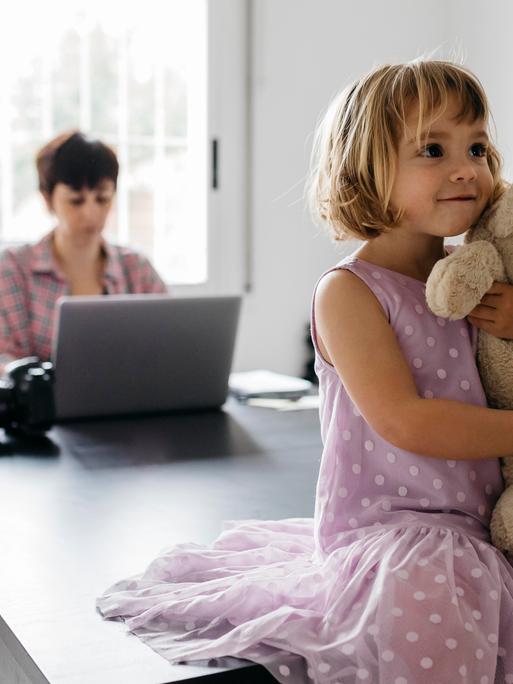 Eine Frau arbeitet an einem Tisch mit dem Laptop. Auf dem Tisch sitzt ein Mädchen, das einen Teddybären im Arm hält.