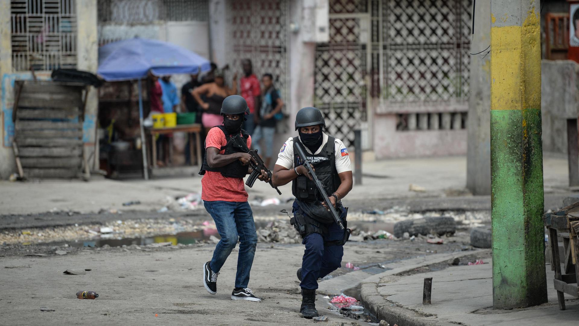 Polizisten gehen mit Gewehr im Anschlag durch Port-au-Prince. Im Hintergrund drängen sich mehrere Menschen in einem Hauseingang.