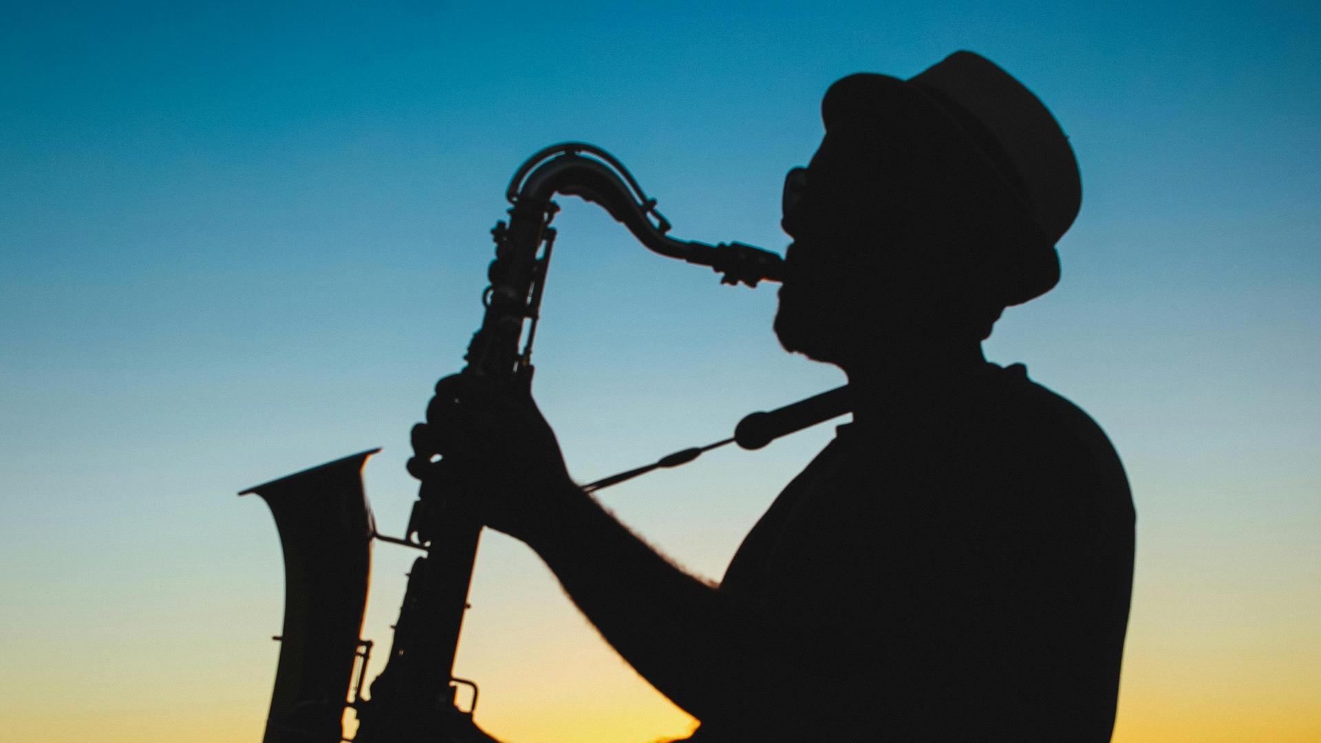 Ein Mann spielt Saxophon vor einem dunkelblau-gold-orangenen Himmel, der die letzten Sonnenstrahlen noch in der Atmosphäre hat. Ein Mann spielt Saxophon vor einem dunkelblau-gold-orangenen Himmel, der die letzten Sonnenstrahlen noch in der Atmosphäre hat.