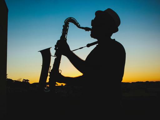 Ein Mann spielt Saxophon vor einem dunkelblau-gold-orangenen Himmel, der die letzten Sonnenstrahlen noch in der Atmosphäre hat.
