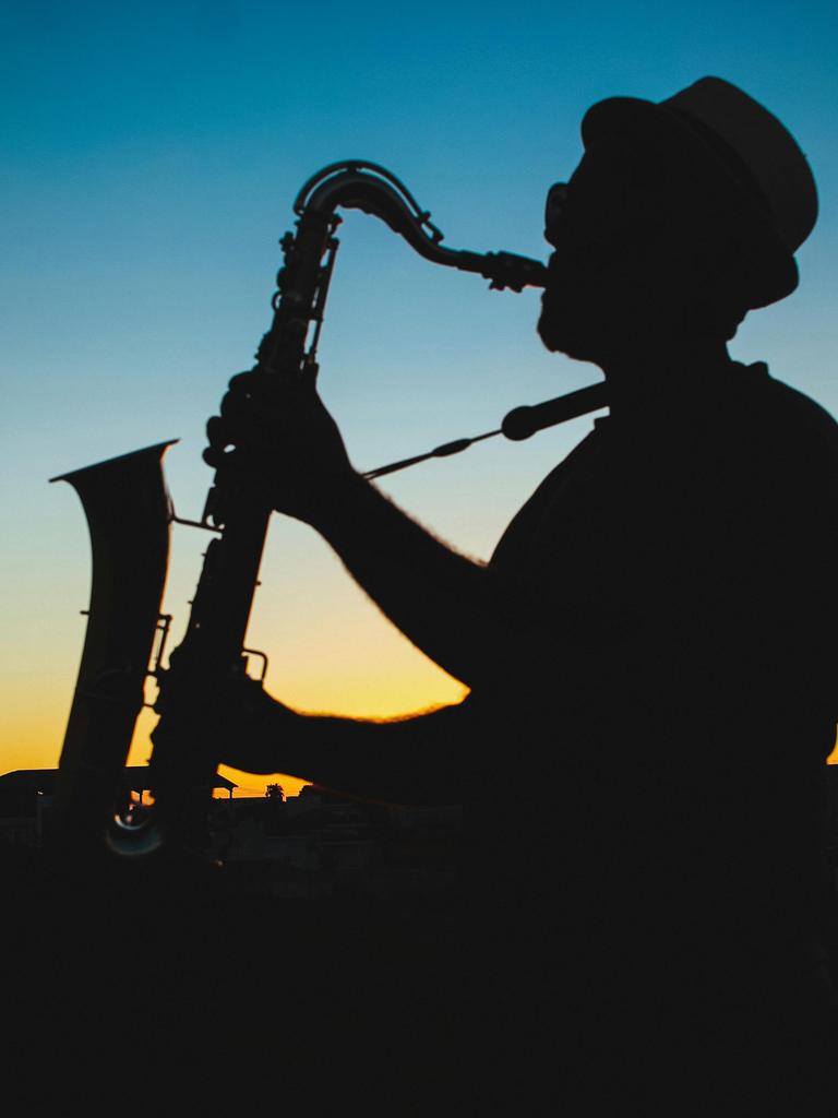 Ein Mann spielt Saxophon vor einem dunkelblau-gold-orangenen Himmel, der die letzten Sonnenstrahlen noch in der Atmosphäre hat.