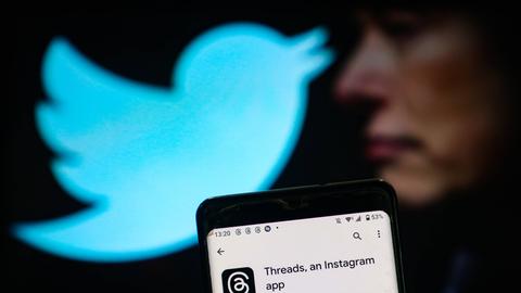 Zu sehen ist die App "Threads" auf einem Handy. Dahinter in der Unschärfe Elon Musk und das Twitter-Logo, der zwitschernde Vogel in hellem Blau.