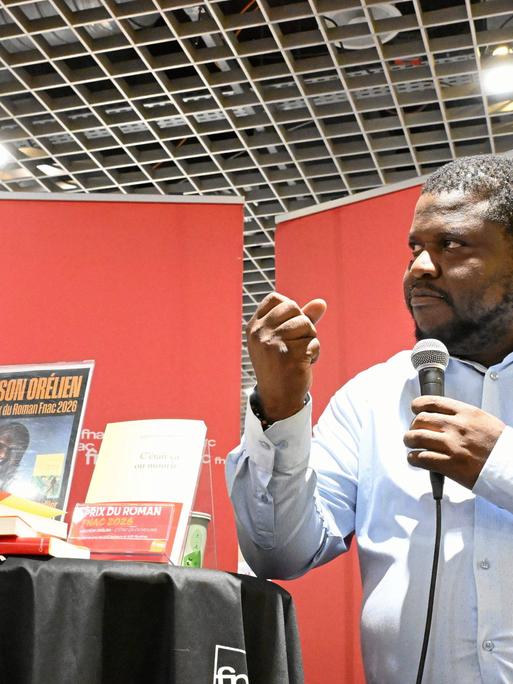 Der kanadisch-haitianische Autor Thélyson Orélien steht mit einem Mikrofon an einem Tisch und ballt eine Faust. Der kanadisch-haitianische Autor Thélyson Orélien steht mit einem Mikrofon an einem Tisch und ballt eine Faust.