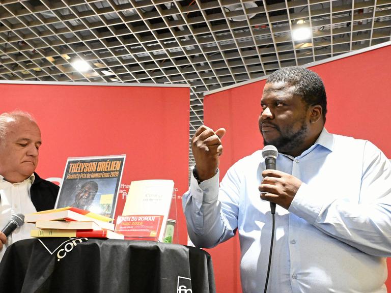 Der kanadisch-haitianische Autor Thélyson Orélien steht mit einem Mikrofon an einem Tisch und ballt eine Faust. Der kanadisch-haitianische Autor Thélyson Orélien steht mit einem Mikrofon an einem Tisch und ballt eine Faust.