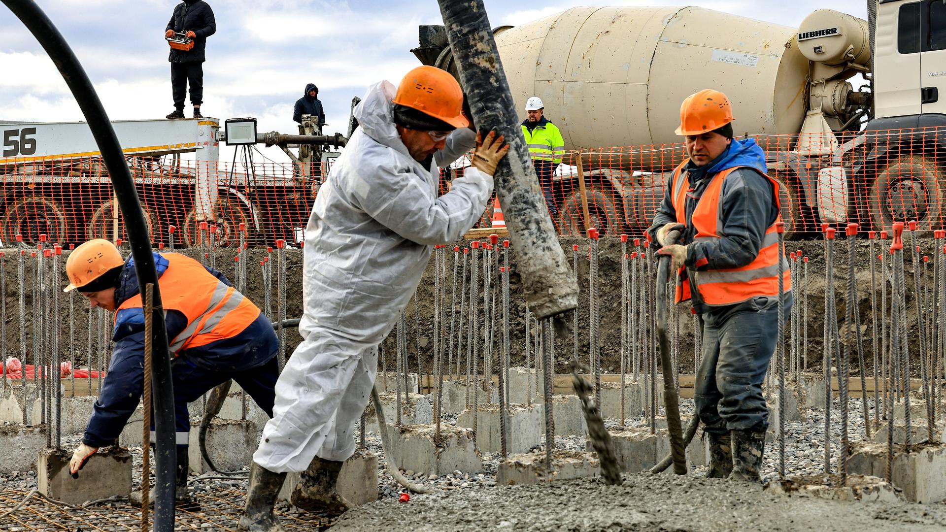 Arbeiter auf einer Baustelle. Im Vordergrund hält ein Mann einen dicken Schlauch aus dem Beton auf ein Stahlgitter gegossen wird. Dahinter stehen weitere Männer und ein Lastwagen.