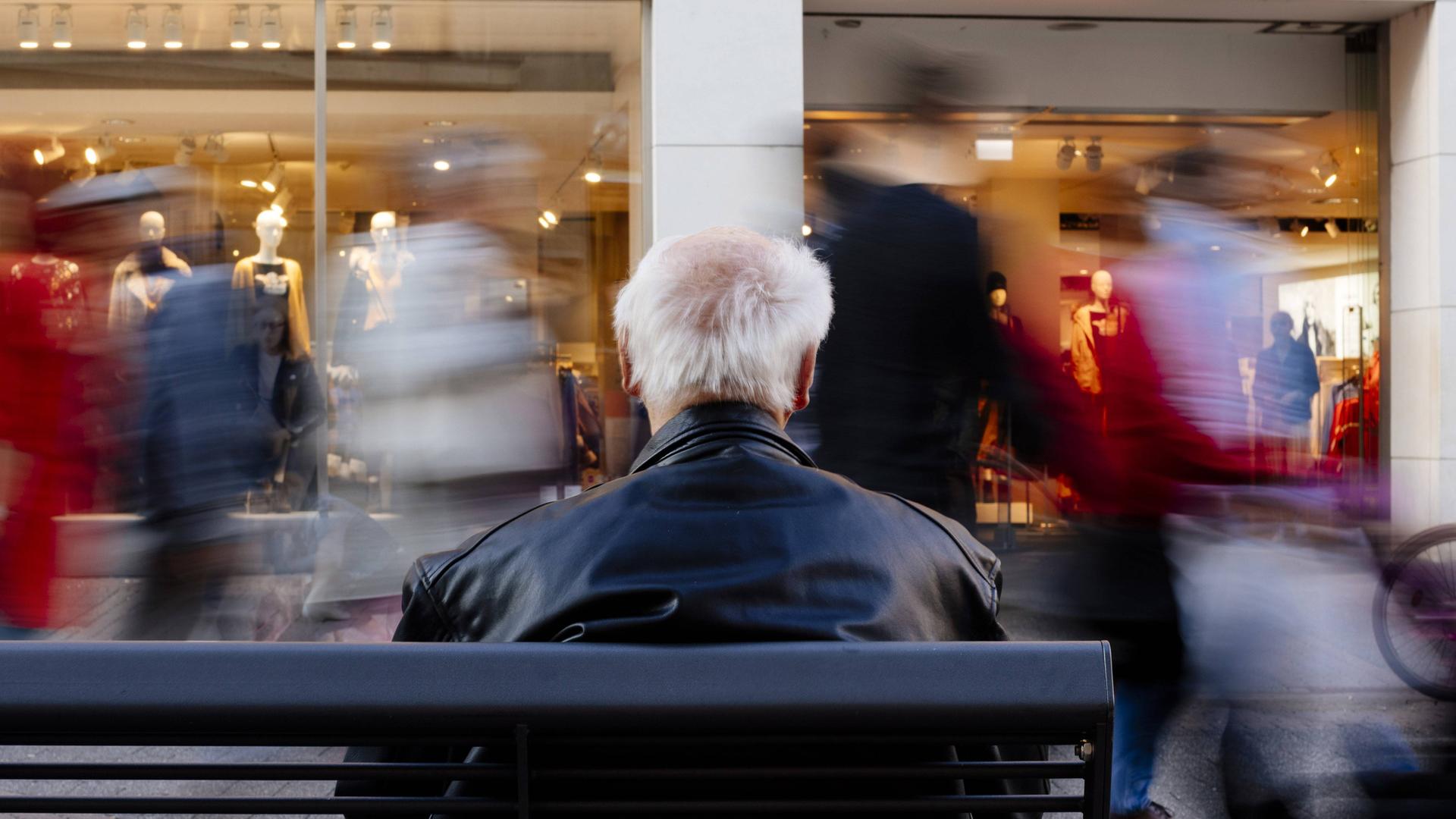 Vor einem älteren Mann, der auf einer Bank in einer Straße sitzt, gehen Menschen vorbei