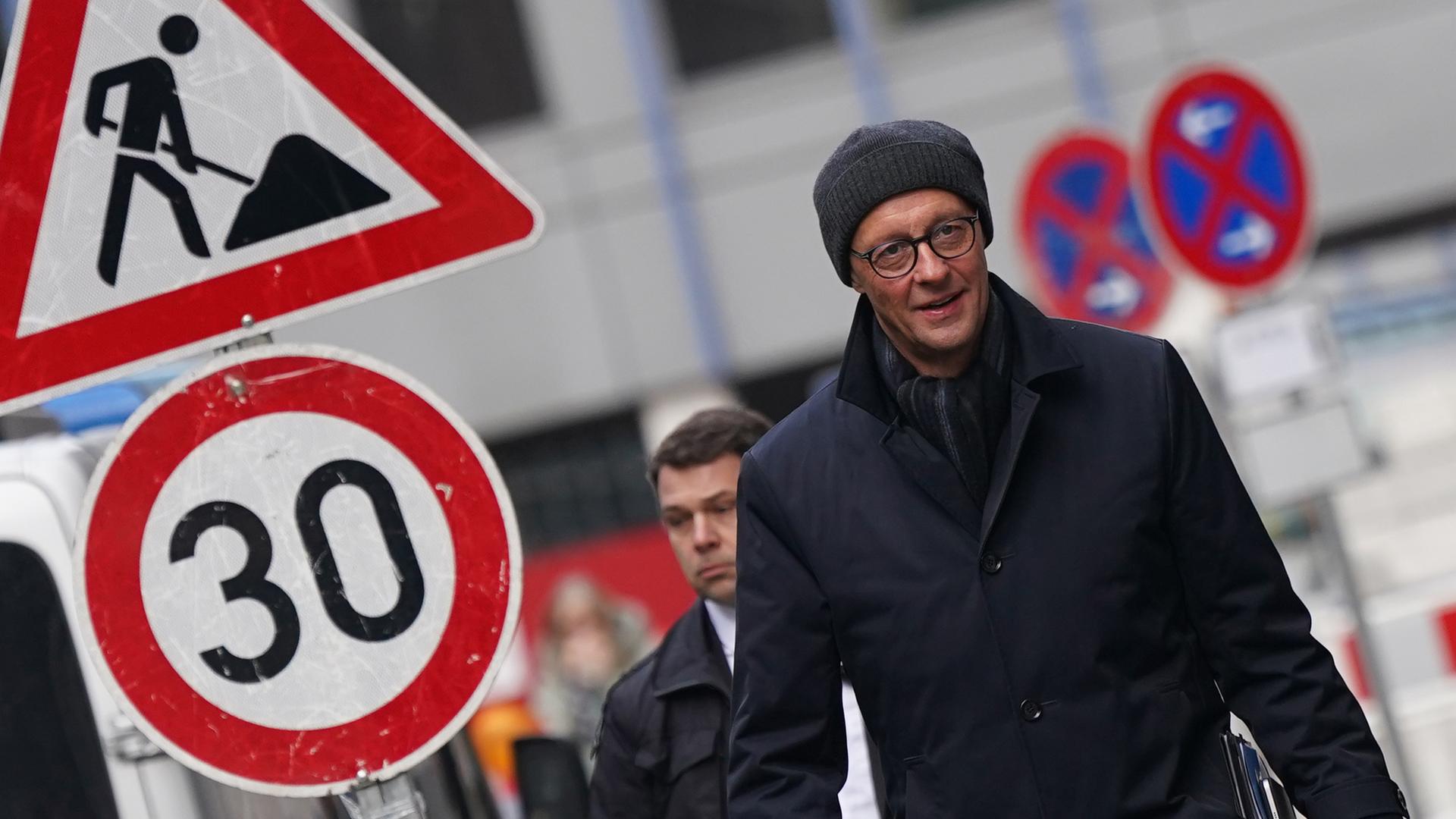 Friedrich Merz, Kanzlerkandidat und Parteivorsitzender der CDU, läuft auf einer winterlichen Straße entlang. Er trägt eine Mütze. Neben ihm ist ein Baustellenschild zu sehen, außerdem ein Schild zur Begrenzung des Tempolimits auf 30 Km/h. 