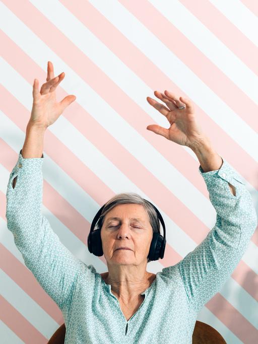 Eine ältere Frau vor einer gestreiften Tapete mit geschlossenen Augen und einem Kopfhörer. Sie streckt beide Arme in die Höhe.